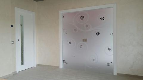 A destra, porta a doppia anta con motivo lavorato in sabbiatura con decori scavati e colorati in nero. A sinistra, striscia di vetro collocata nella nicchia della porta in legno, il cui decoro richiama quello dell infisso accanto.