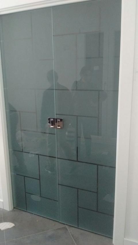 Porta in vetro temperato 8mm trasparente decori rettangolari, disposti in varie angolazioni, lavorati in sabbiatura, lasciando uno spazio fra le figure geomentriche di 1cm in trasparenza. In seguito è stato applicato su tutta la superficie un prodotto acidante per rendere satinata la parte in sabbiatura. Maniglie e accessori in acciaio.