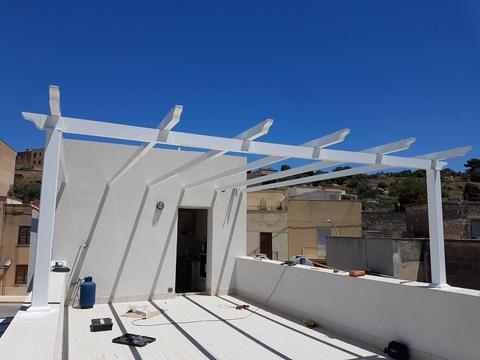 Pergolato tradizionale in alluminio anodizzato bianco opaco inserito nella terrazza di un B&B. La struttura è composta da travi ancorate su un muro portante e piantoni.
